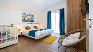 A modern hotel room with a wooden floor, a bed with white bedding and orange pillows, a colorful crib, a wicker chair, and a large window with blue curtains. - Amazing home in Pridraga (Pridraga)