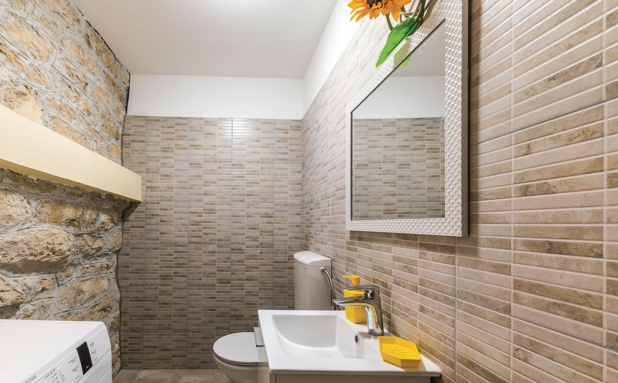 Un baño con inodoro, lavamanos y espejo. Las paredes están cubiertas de azulejos con diseño de ladrillo, y hay una pared de piedra en un lateral.