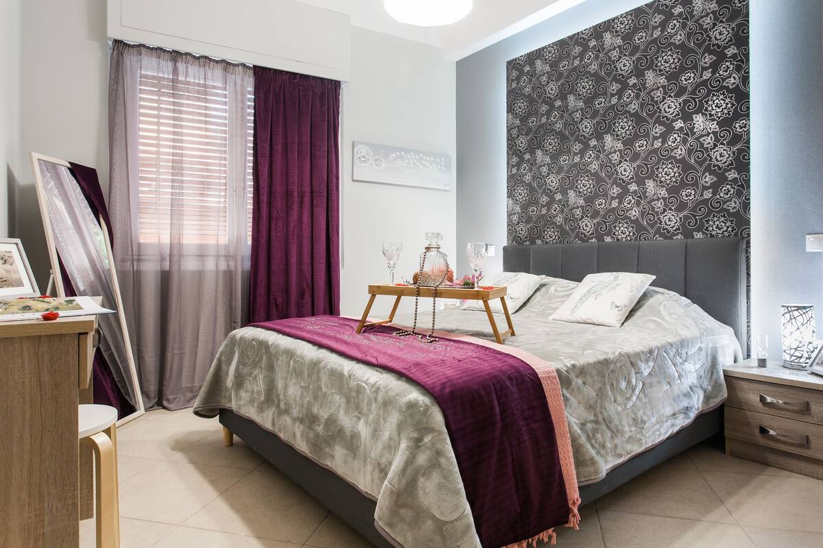 Ein Schlafzimmer mit einem Bett, einem Nachttisch, Vorhängen und einer Wand mit einem floralen Muster.