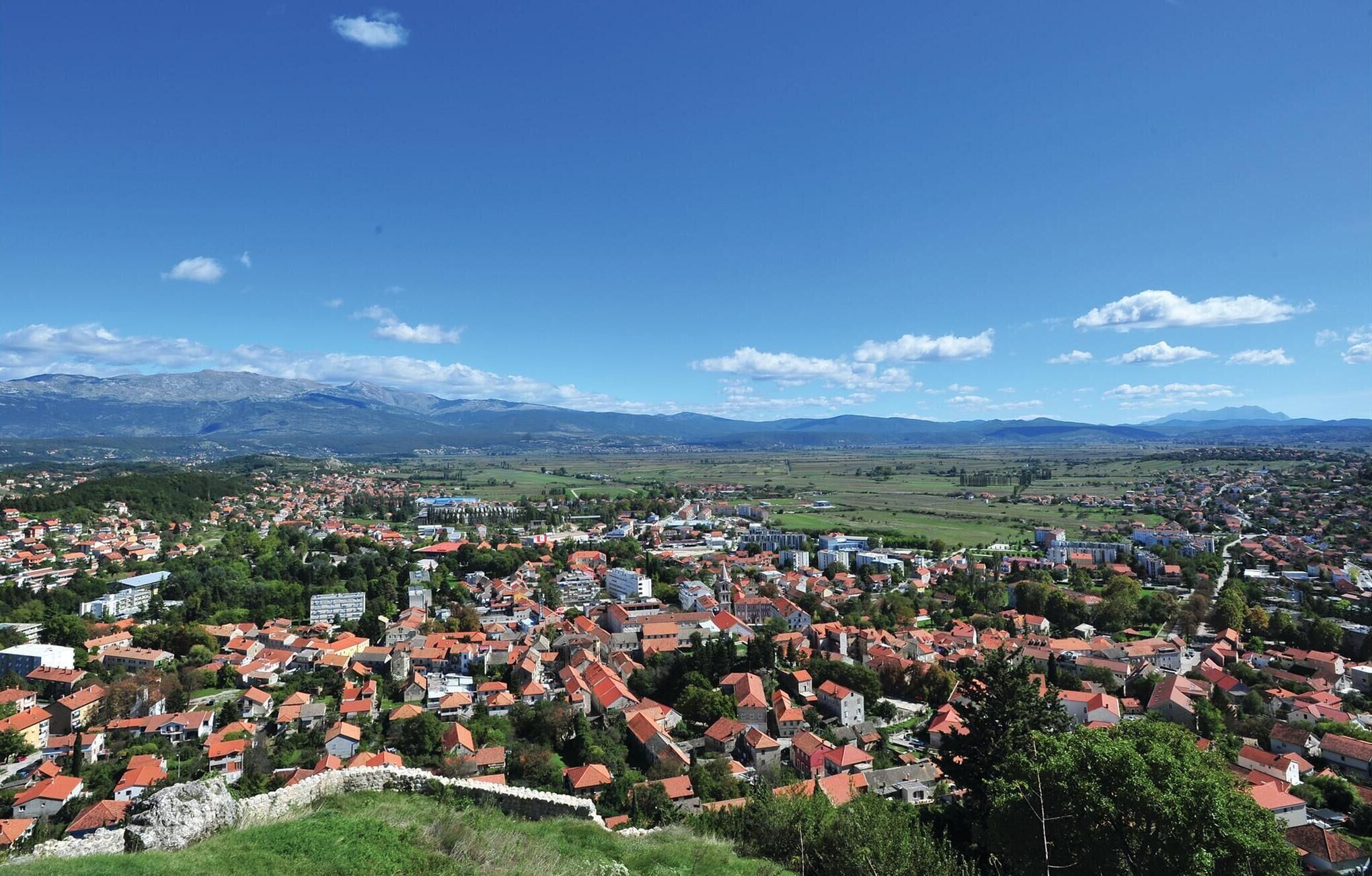 A town with red-roofed houses, a stone wall, and mountains in the background.