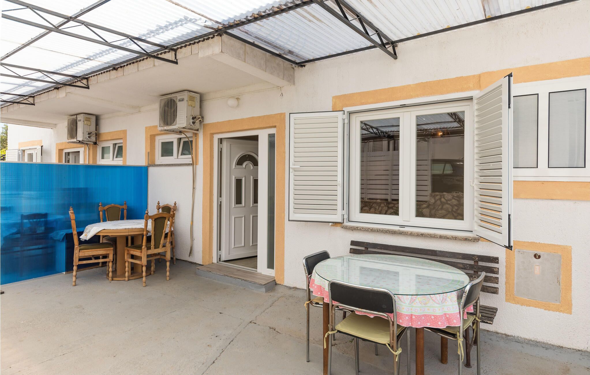 Une terrasse extérieure couverte, avec une table et des chaises, une clôture bleue et une porte donnant sur l’intérieur.