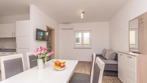 Une cuisine compacte avec des meubles blancs, une table à manger entourée de chaises grises à coussin, un panier de fruits et un vase rempli de fleurs.