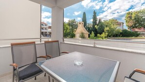Un balcon avec une table et des chaises, donnant sur une rue bordée d’immeubles et d’arbres.