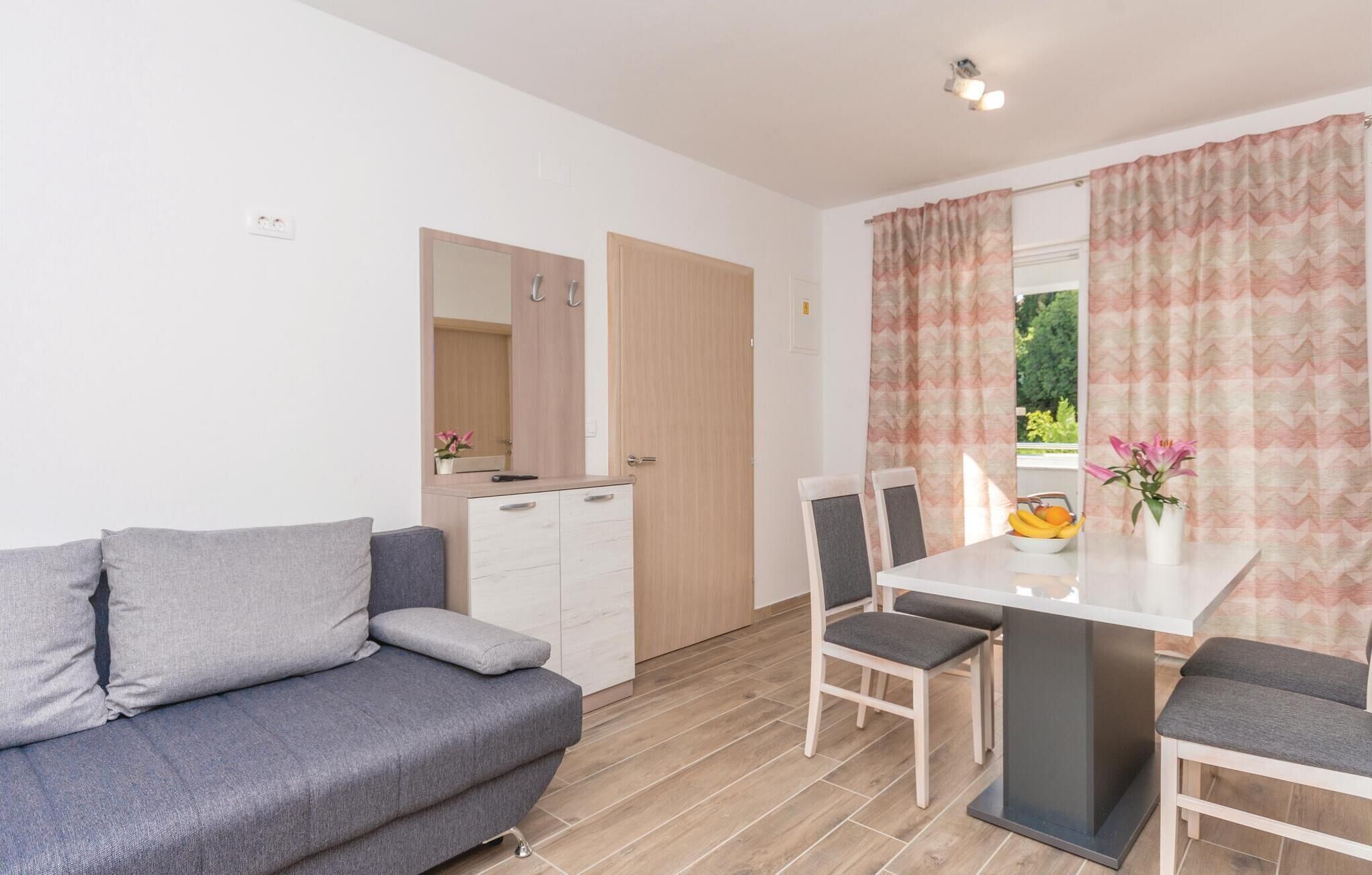 Ein modernes Zimmer mit einem grauen Sofa, einem weißen Esstisch mit zwei Stühlen, einer kleinen Kommode und einem Fenster mit gemusterten Vorhängen.
