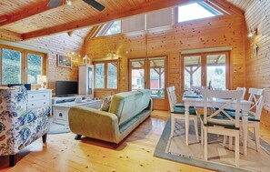 Cozy cabin-style room with a high ceiling and wooden beams, featuring a sofa, dining table, and TV.