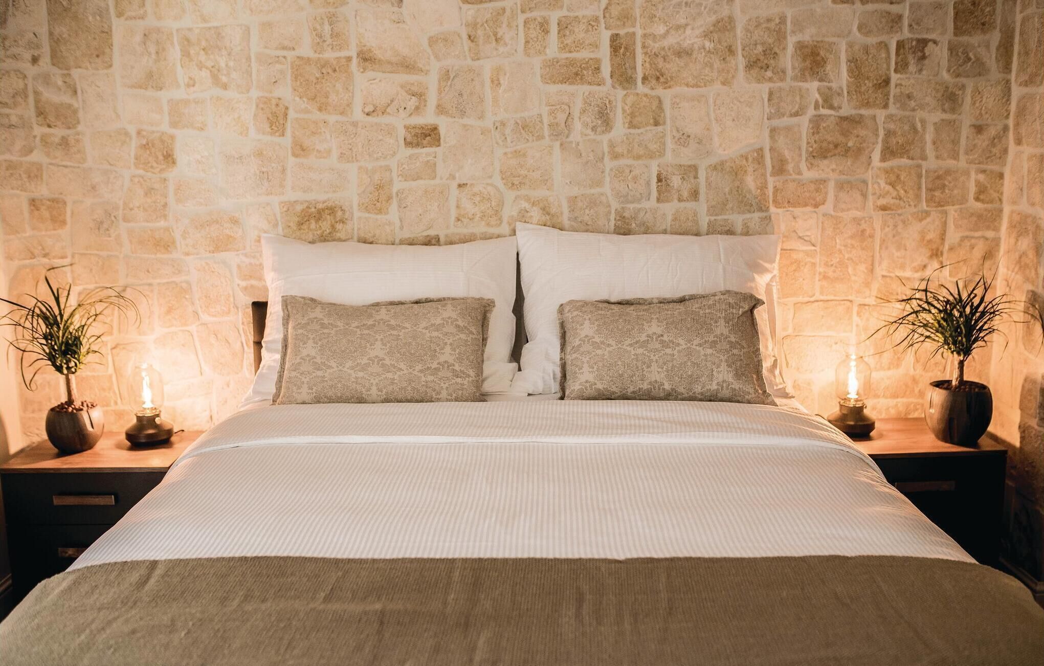 Una camera da letto con parete in pietra, un letto con biancheria bianca, due comodini con lampade e due piante in vaso.
