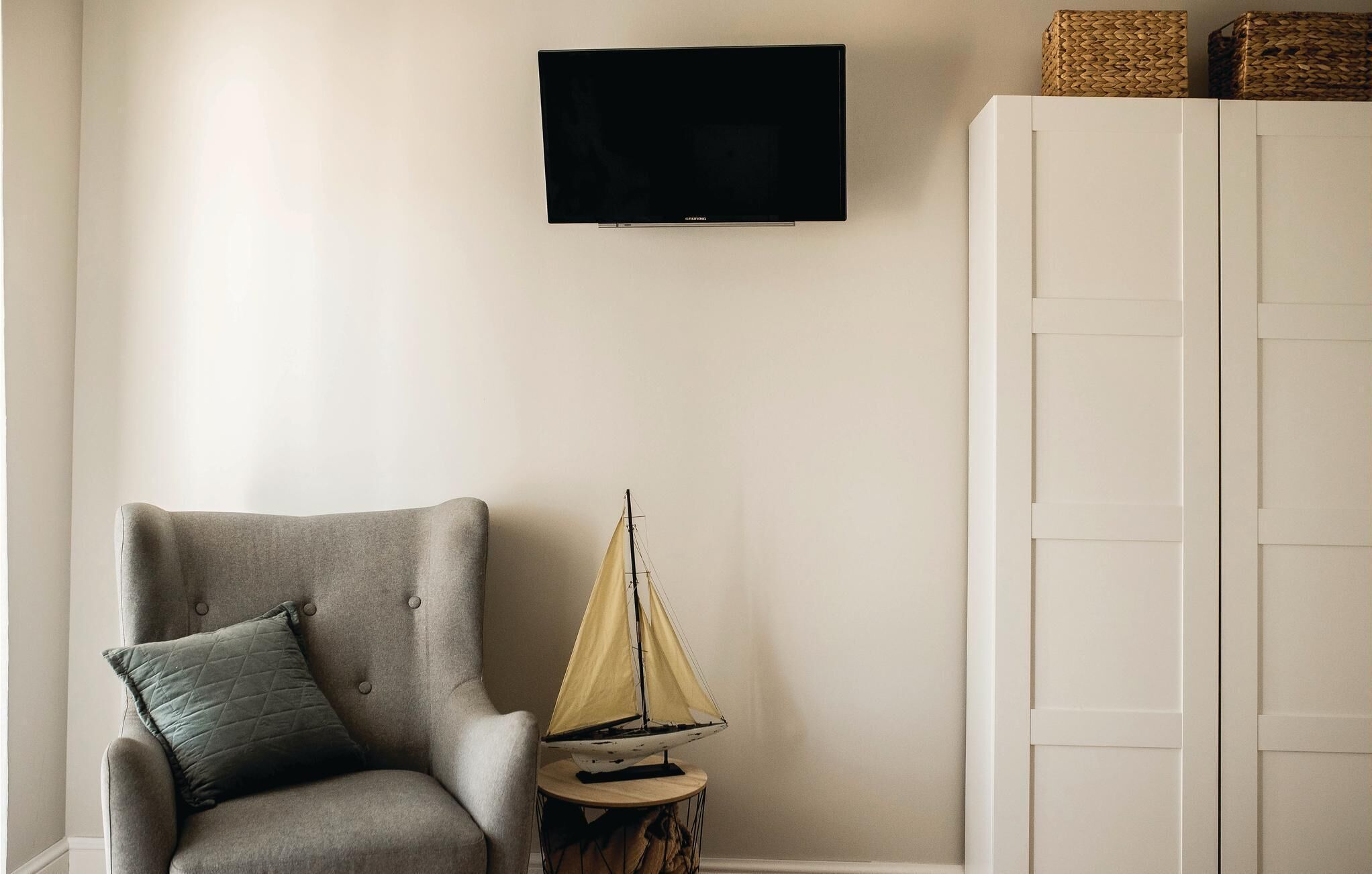 Una habitació amb una armchair gris, una taula rodona petita amb un model de veler, una televisió a la paret i un armari blanc.
