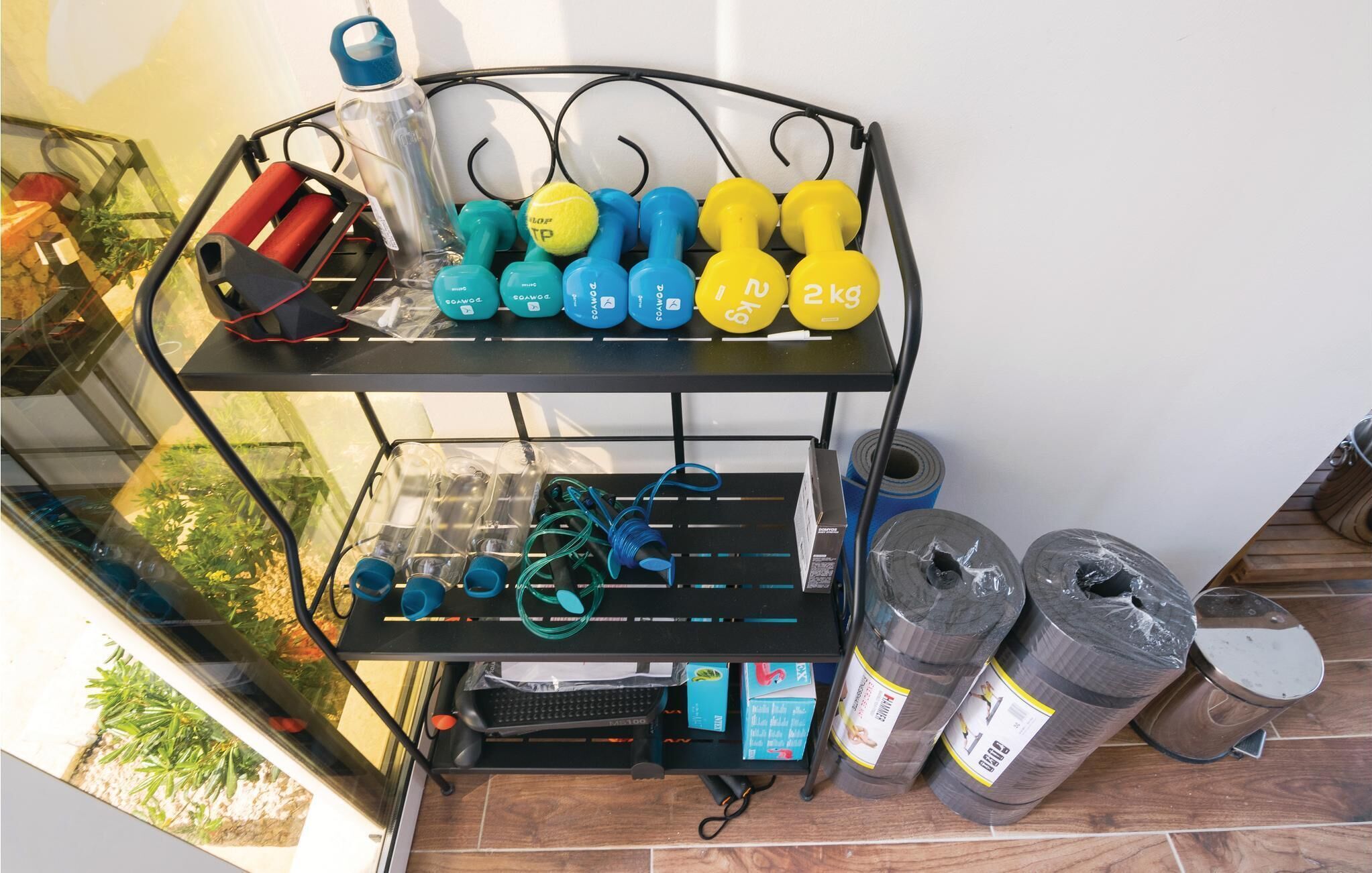 Een metalen rek met fitnessapparatuur, waaronder gewichten en weerstandsbanden, op een balkon met uitzicht op groen.