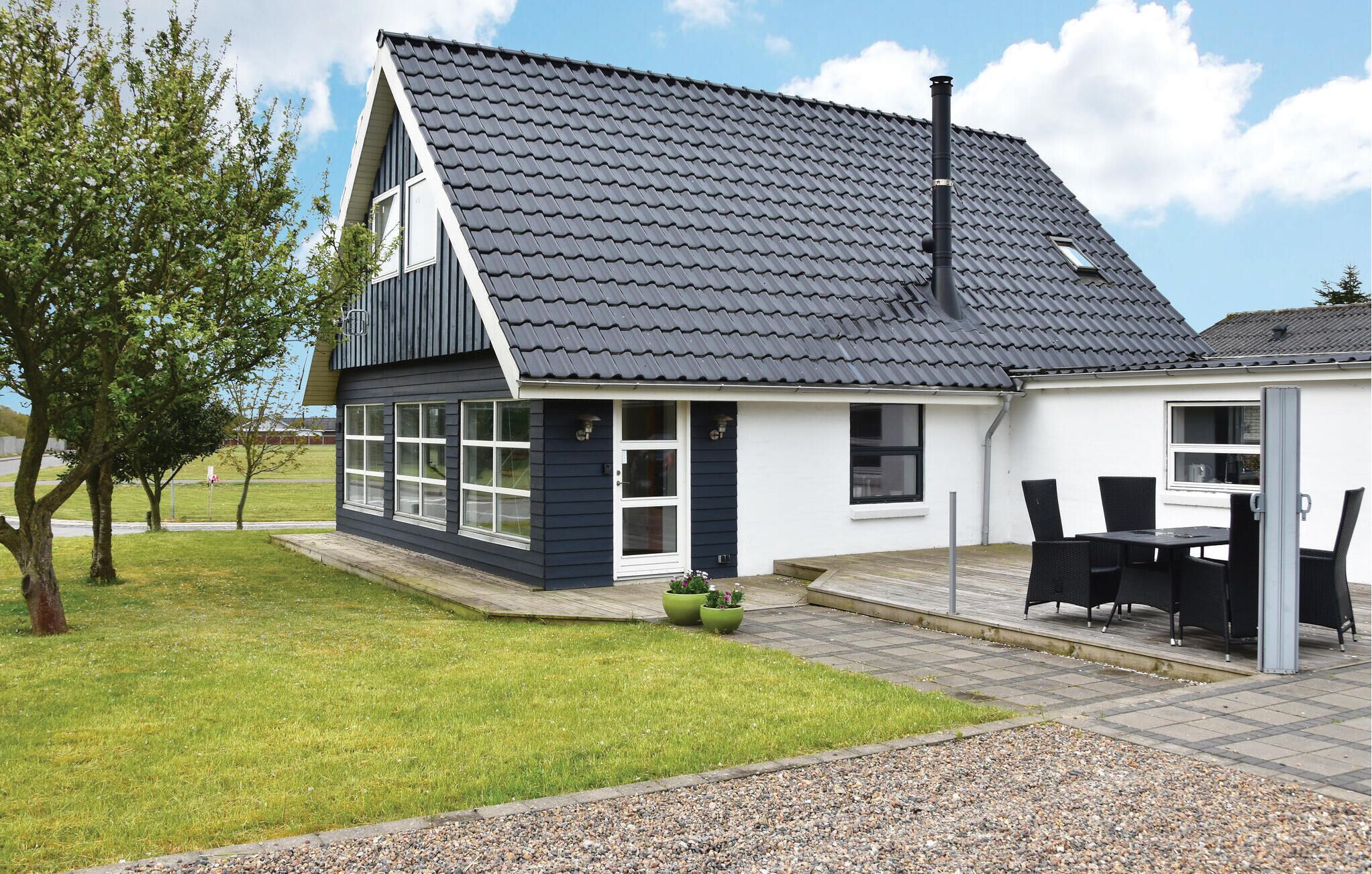 Une maison moderne avec un toit foncé, de grandes fenêtres et un patio aménagé avec du mobilier d’extérieur.