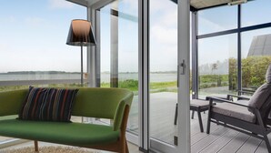 A modern room with a green sofa, a striped pillow, and a lamp. It features large glass doors leading to an outdoor deck with a view of a lake and greenery.