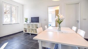 Un comedor moderno con una mesa y sillas blancas, un televisor sobre un soporte blanco y un jarrón con flores.