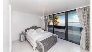 Quarto com cama, mesa de cabeceira, lustre e vista para o mar através de amplas portas de vidro.