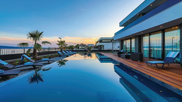 Piscina infinita no terraço com espreguiçadeiras e deck de madeira, com vista para a paisagem urbana ao pôr do sol.