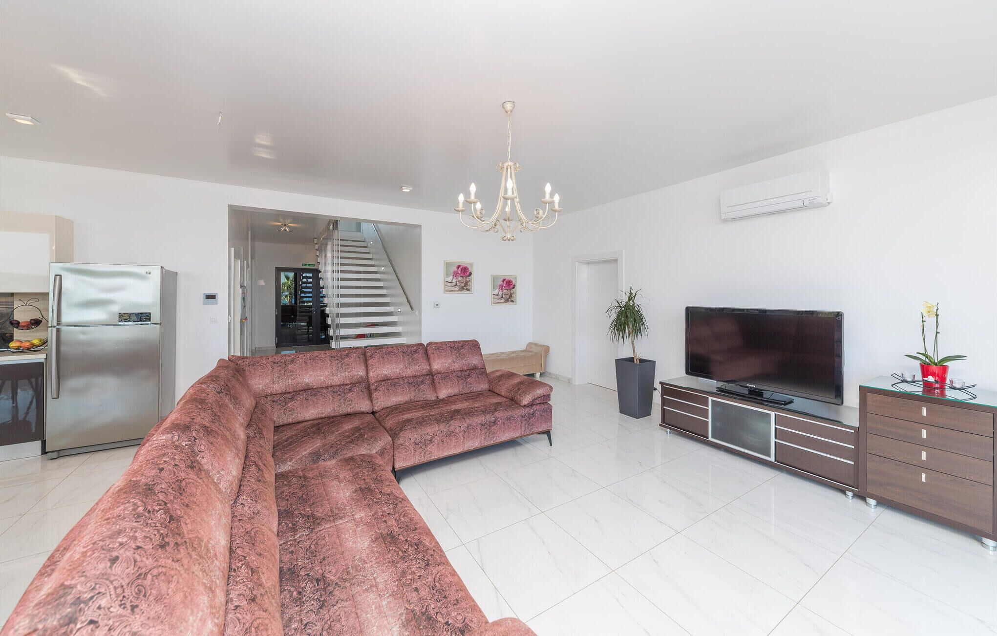 A modern living room with a sectional sofa, a flat-screen TV, a dining area with a refrigerator, and a staircase leading to an upper level.