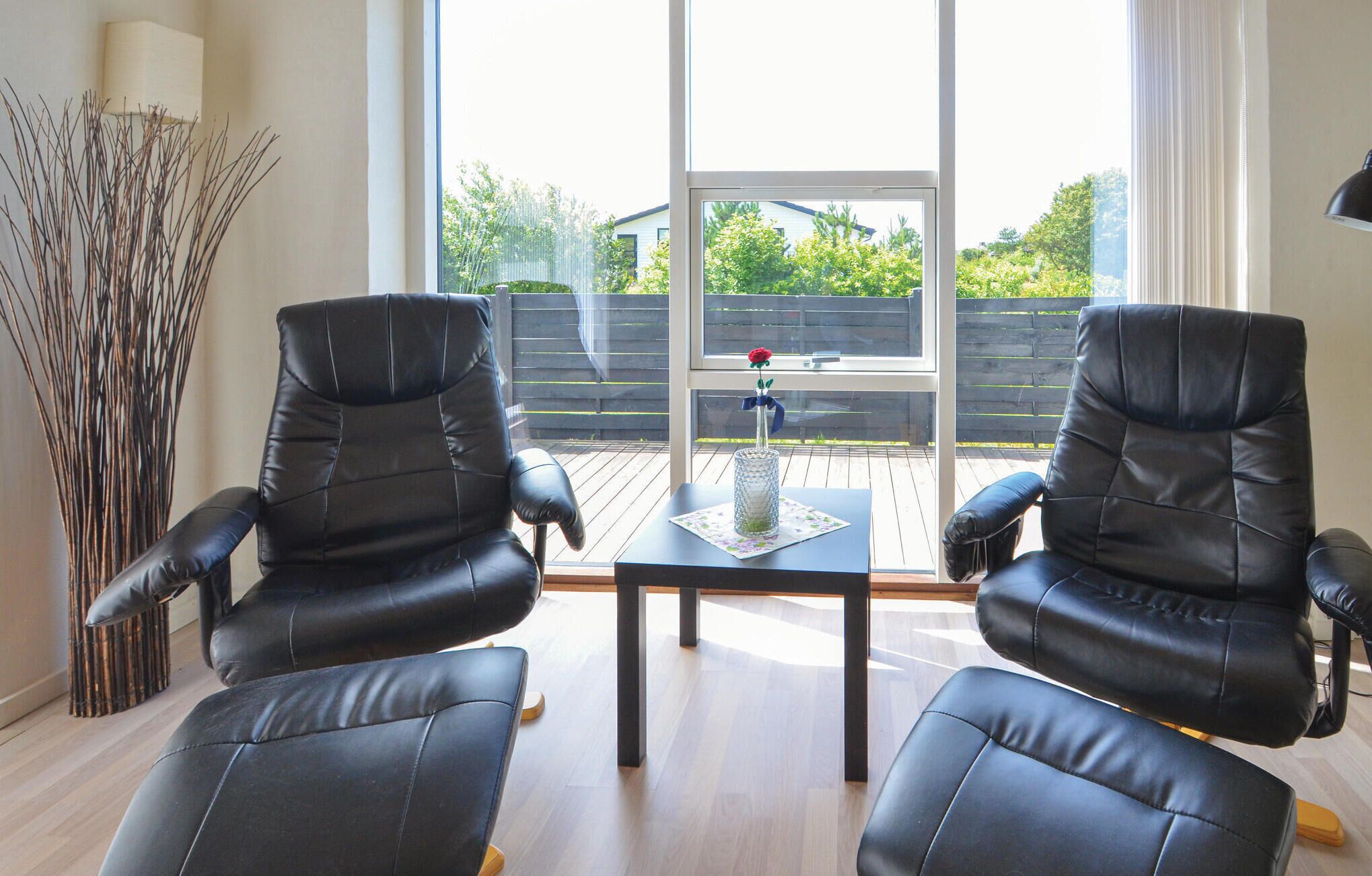 A modern living room with two black leather chairs facing a glass table. A large window offers a view of a wooden deck and greenery outside.