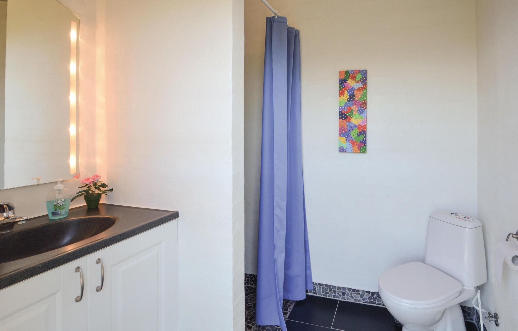 A bathroom with a white toilet, a sink with a mirror, a blue shower curtain, and a colorful abstract painting on the wall.