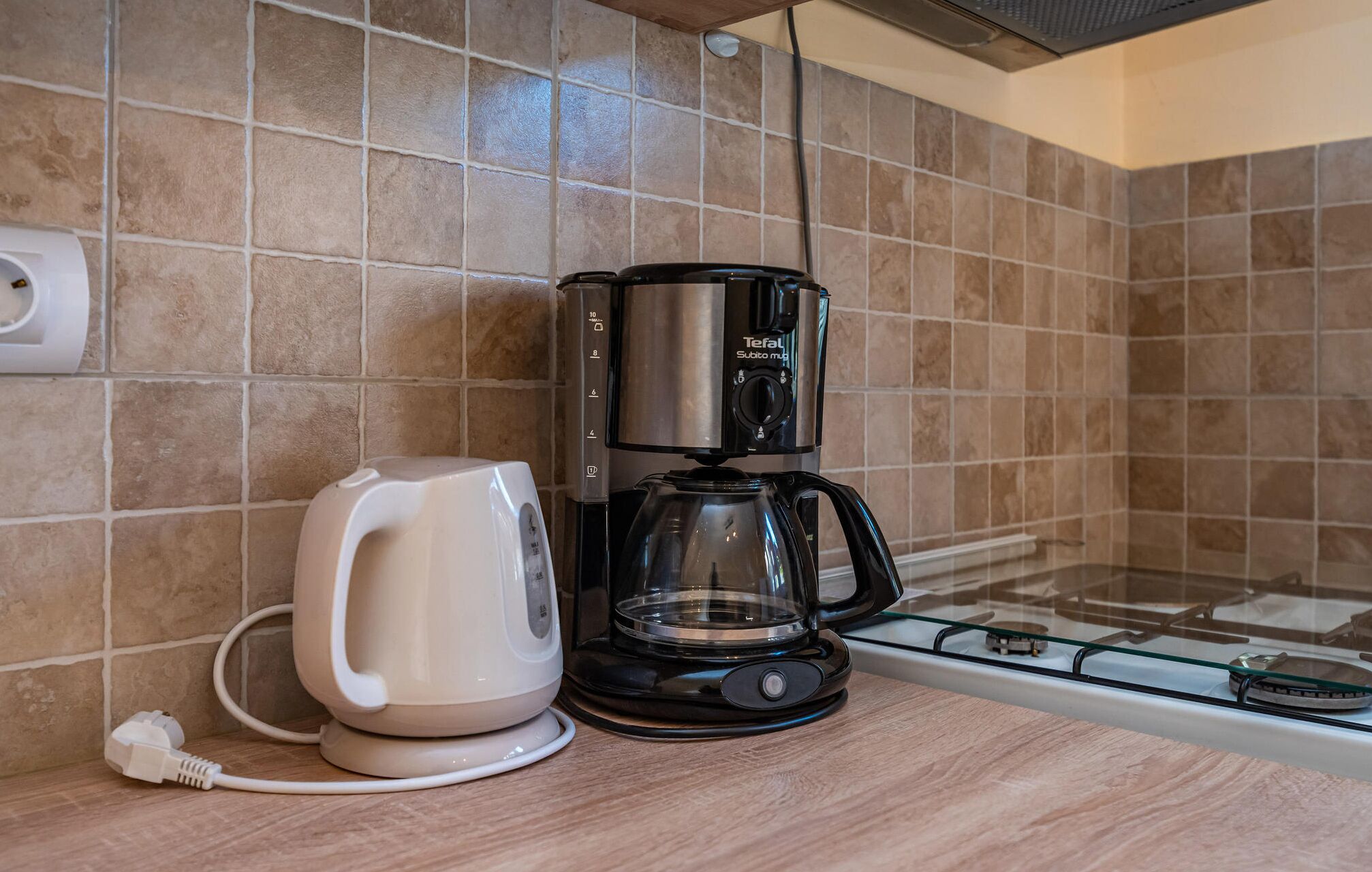 Kaffemaskine og elkedel på en køkkenbordplade med flisebeklædt bagvæg.