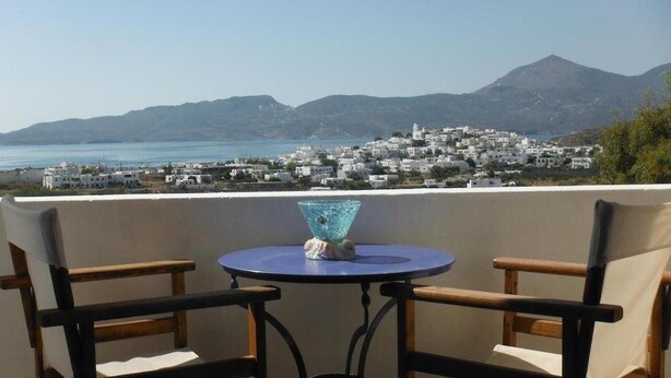 A balcony with two chairs and a table, overlooking a coastal town with white buildings and a mountain in the background. - Kapetan Giannis (Milos)
