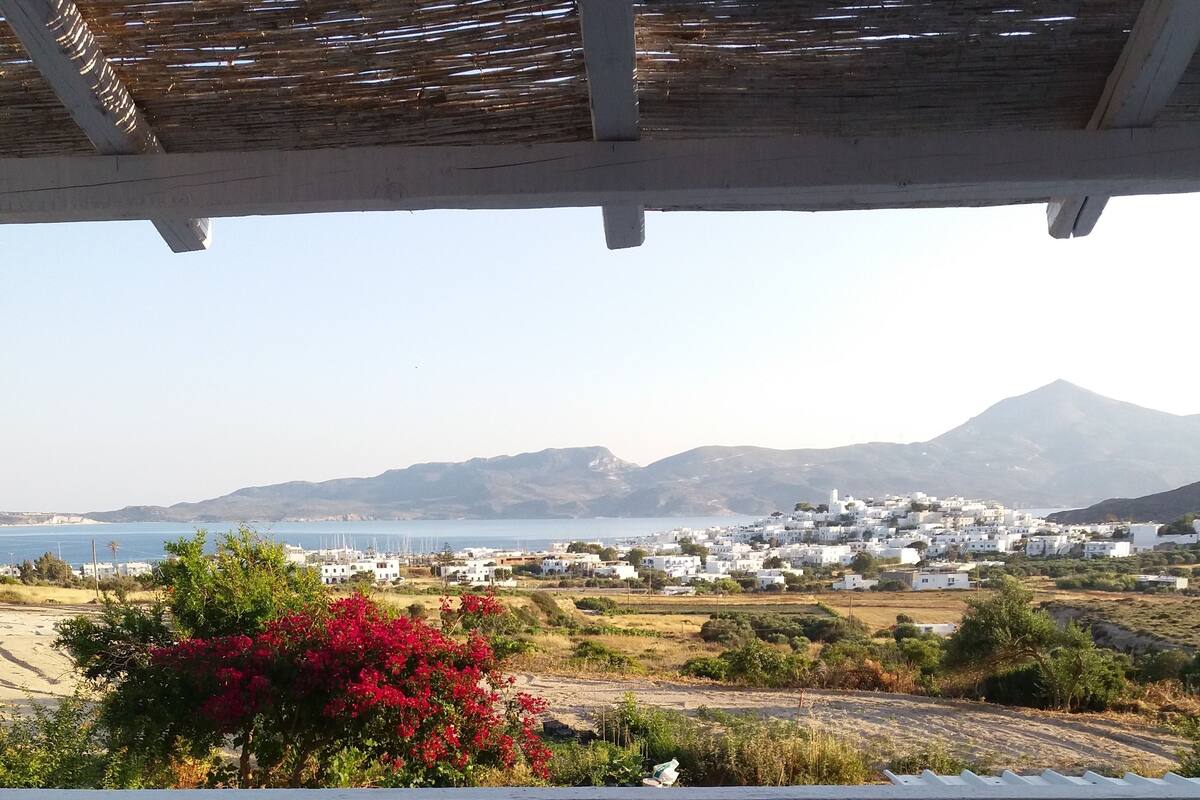 A terrace with two wooden chairs and a small round table, overlooking a coastal landscape with white buildings and a mountain in the distance.