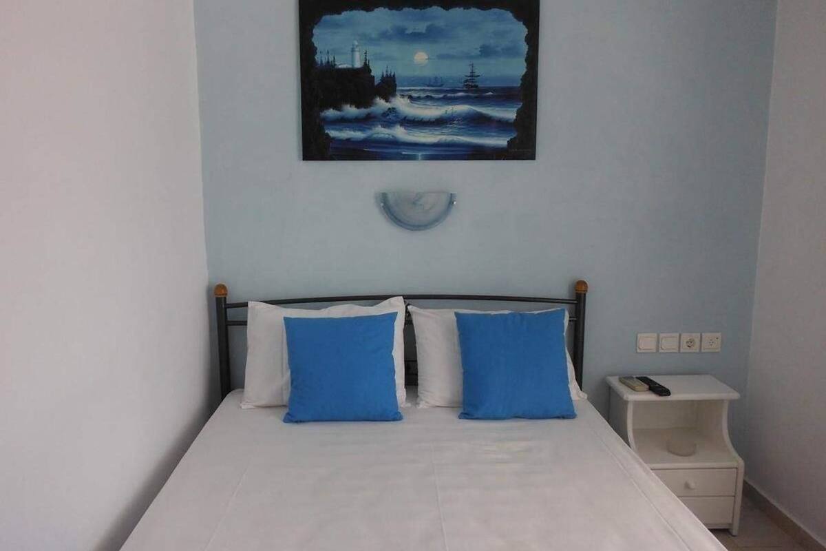 A bed with white and blue bedding, a painting of a seascape above it, and a bedside table with a remote control.