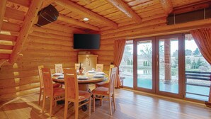 A wooden interior with a dining area, a flat-screen TV mounted on the wall, and a large sliding glass door leading to an outdoor area.