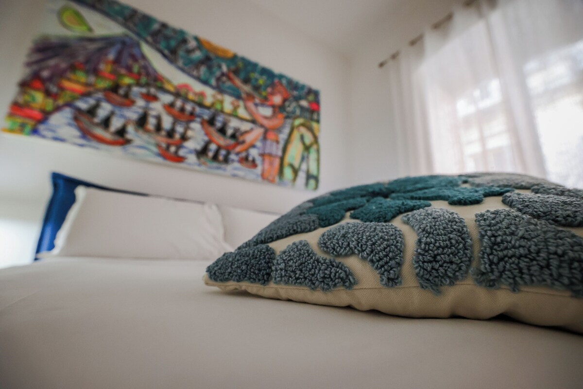 A bed with a white pillow and a textured blue pillow, a colorful abstract painting on the wall, and a window with white curtains.