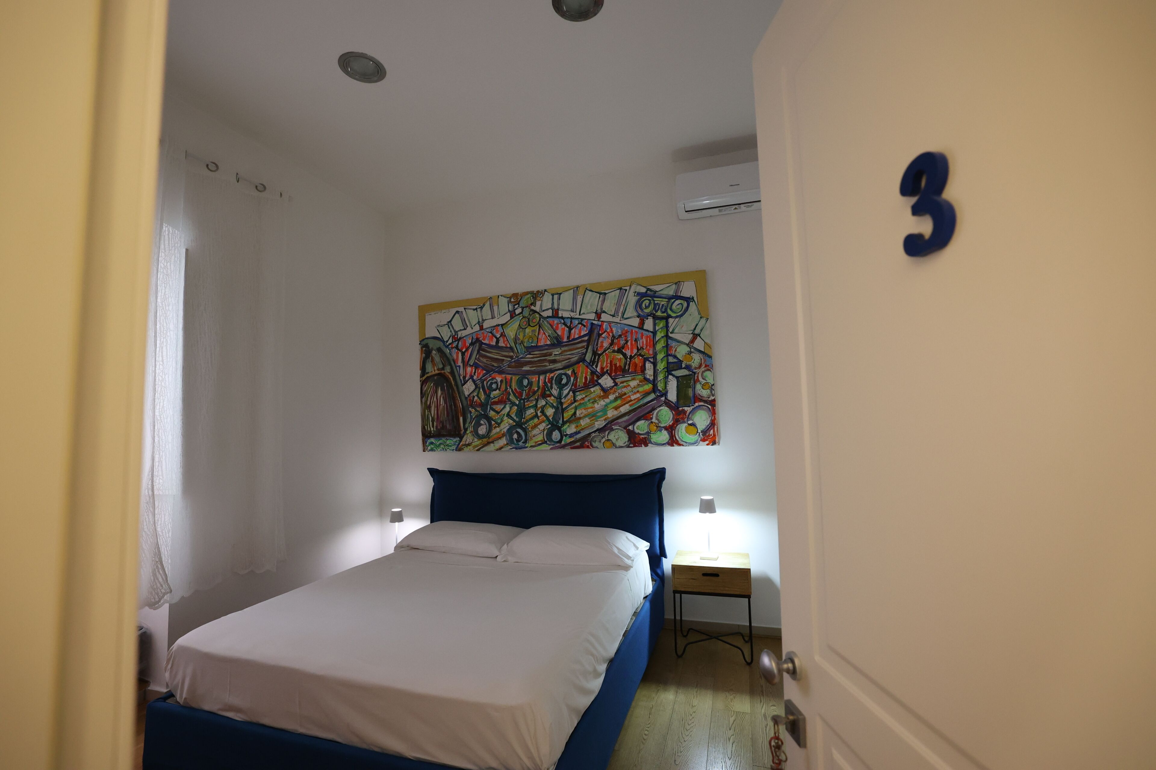 A bedroom with a bed, a nightstand, a wall-mounted painting, and a door with the number 3.