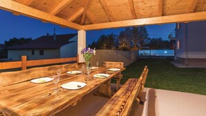 An outdoor wooden deck with a dining table set for four, surrounded by a wooden fence, and a view of a swimming pool and houses in the background. - Lovely home in Pristeg (Pristeg)