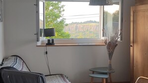 Una stanza con una grande finestra, un soffitto in legno, una poltrona a dondolo, un tavolino con un vaso e un letto con biancheria a motivi.