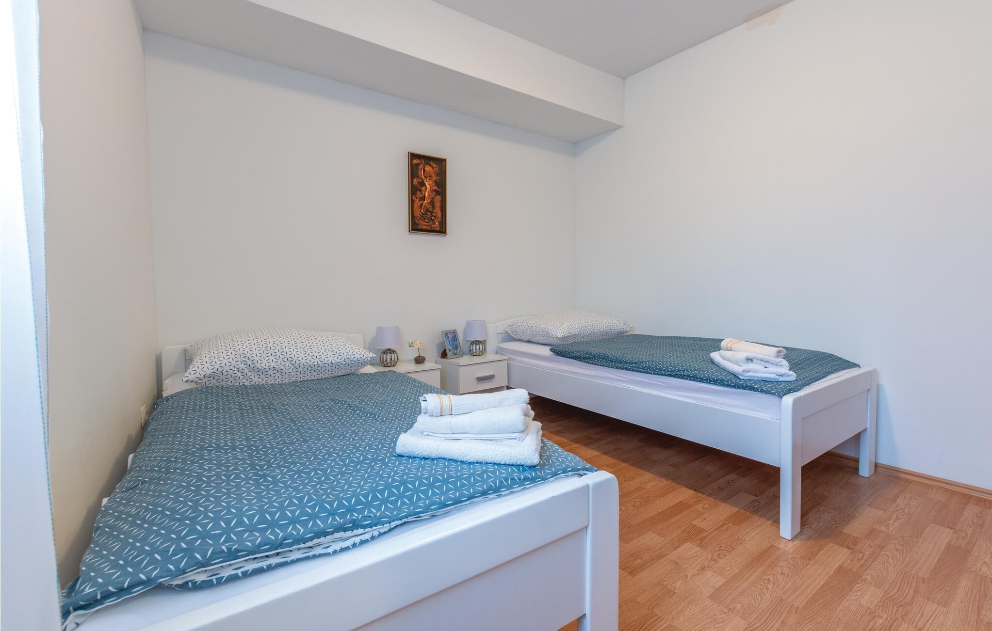 A bedroom with two beds, each with a patterned bedspread and pillows, a bedside table with a lamp, and a framed picture on the wall.