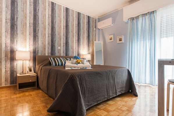 A bedroom with a wooden floor, a bed with a dark bedspread, a nightstand, a lamp, a mirror, and a window with curtains.