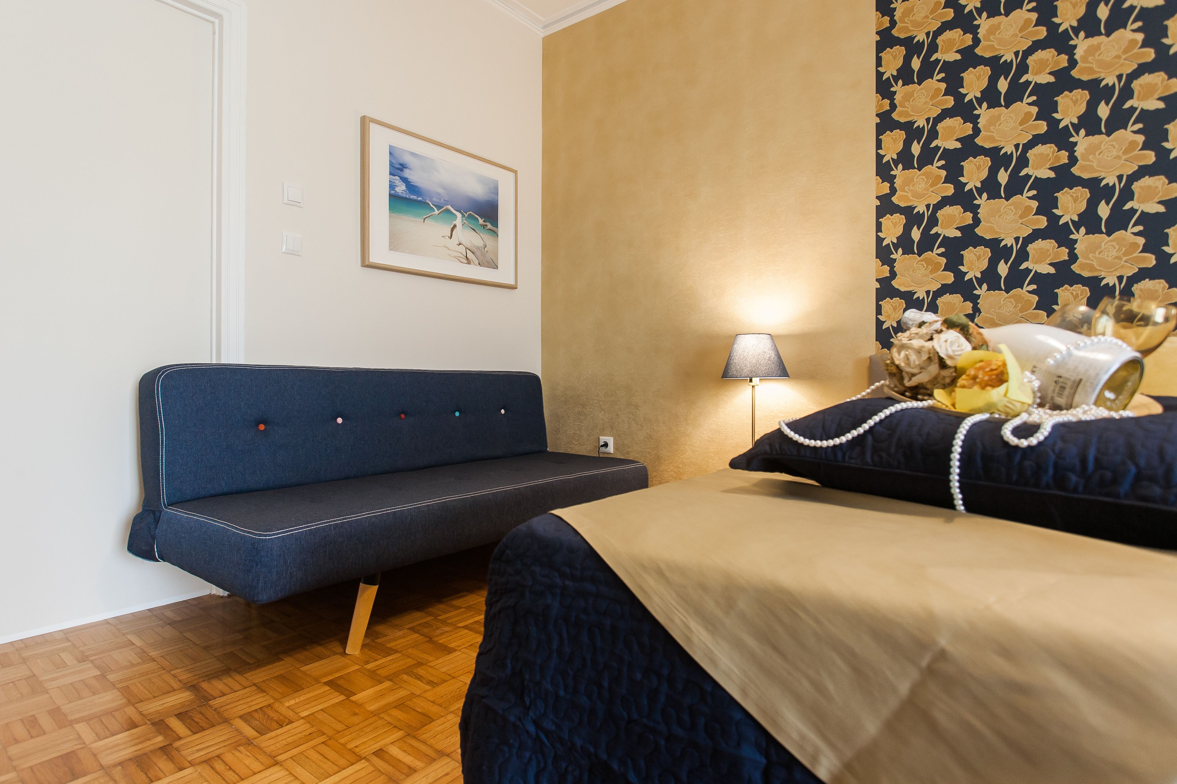 Une pièce comprenant un canapé, un lit recouvert d’une couverture bleue, une lampe et un mur orné d’un motif floral.