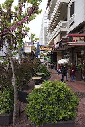 An outdoor seating area with tables and chairs, surrounded by greenery and a building with shops.