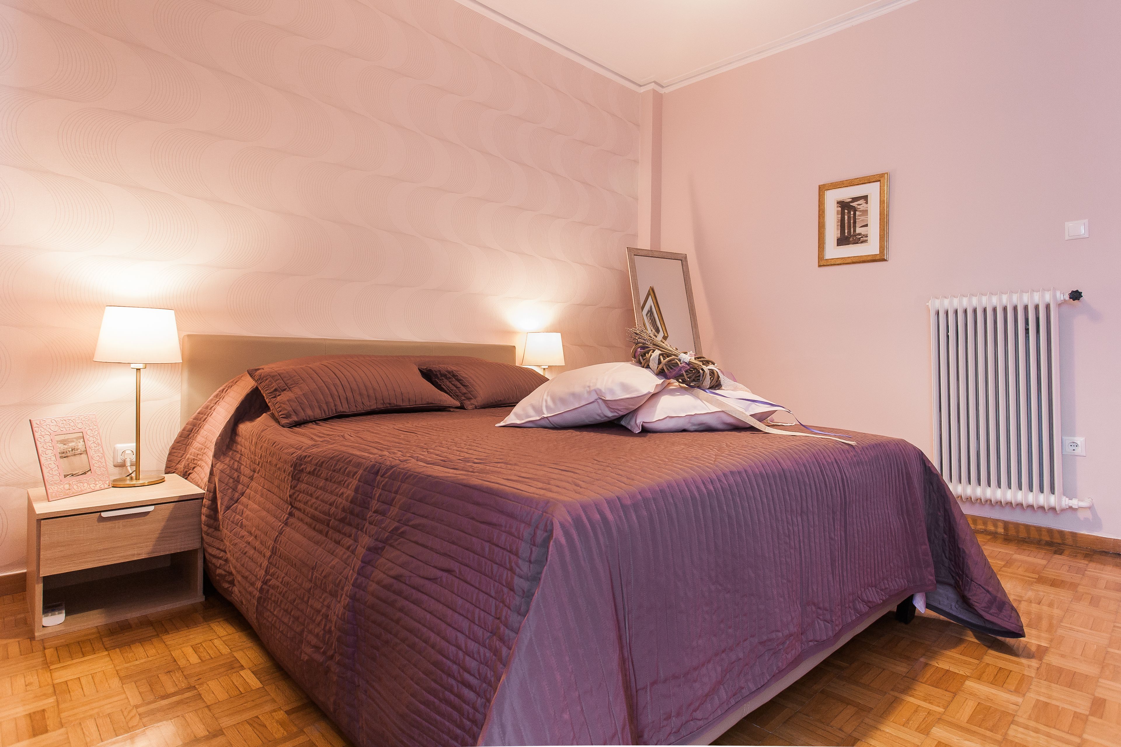A neatly made bed with a purple bedspread and pillows, a bedside table with a lamp, a framed picture on the wall, and a radiator.