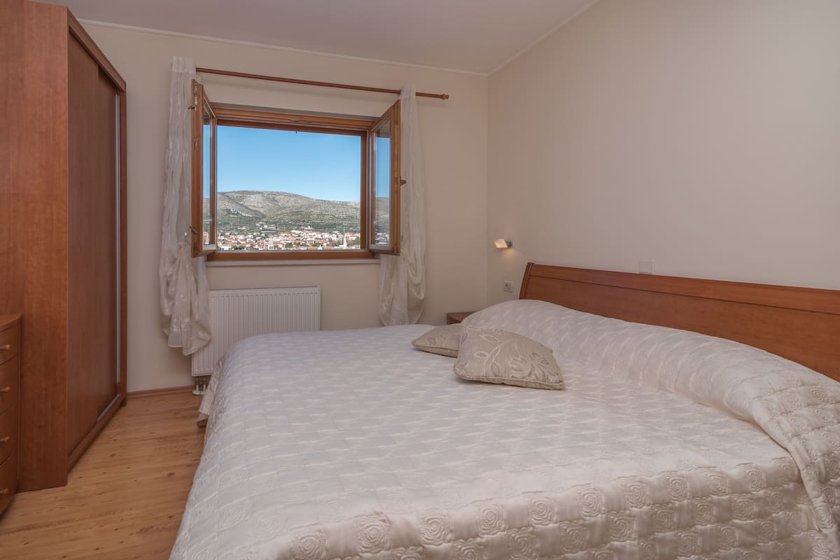 A bedroom with a wooden headboard, a large bed with white bedding, a window with a view of a town and hills, and a radiator under the window.