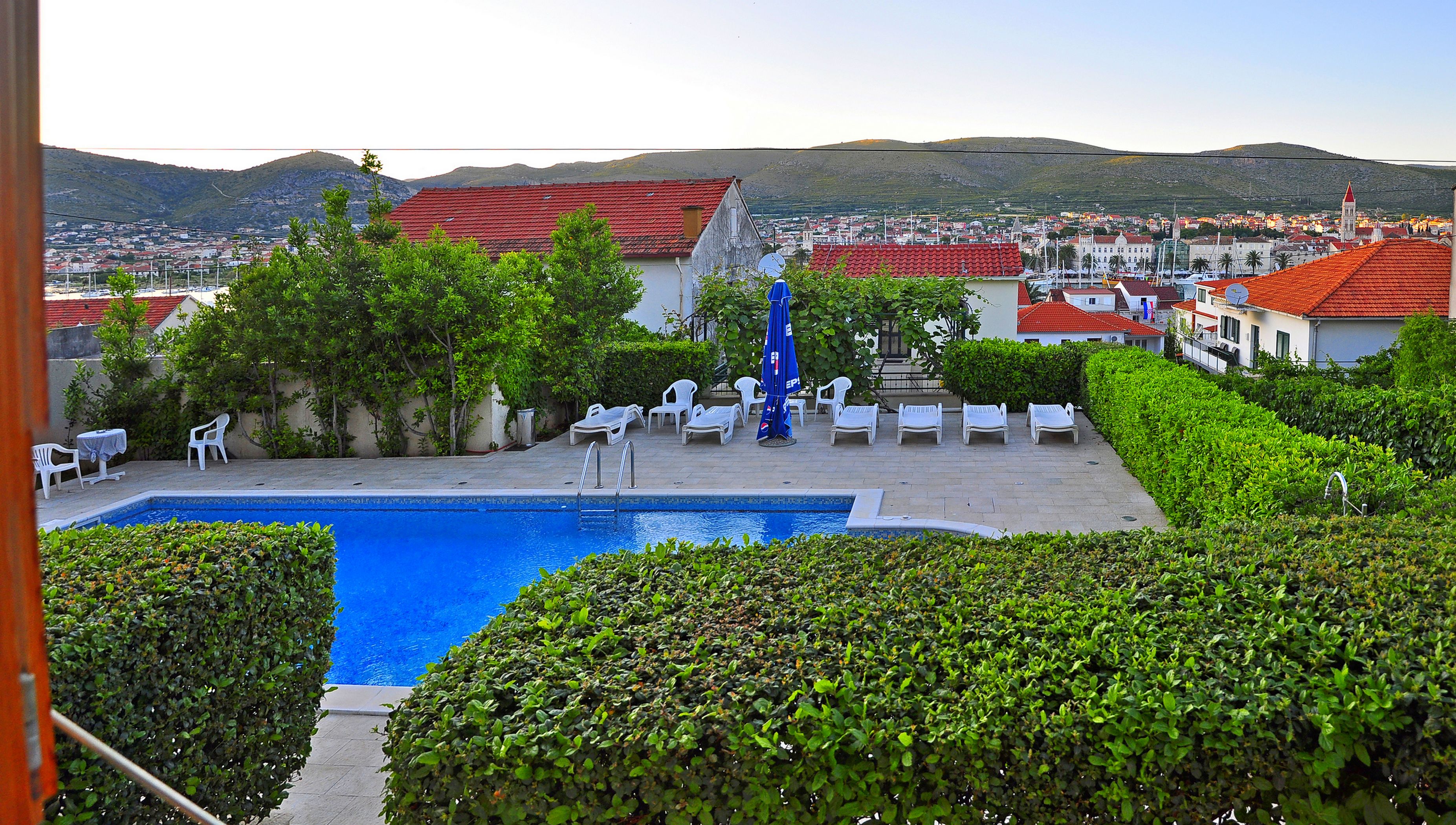 Ein Pool mit Liegestühlen und Blick auf eine Stadt mit roten Dächern sowie einen Hügel im Hintergrund.