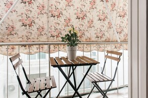 Un balcon agrémenté d’un rideau à motifs floraux, d’une table en bois et de deux chaises en bois.