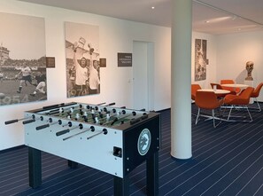 Una habitación con mesa de fútbol, comedor con sillas anaranjadas y una pared decorada con recuerdos deportivos.