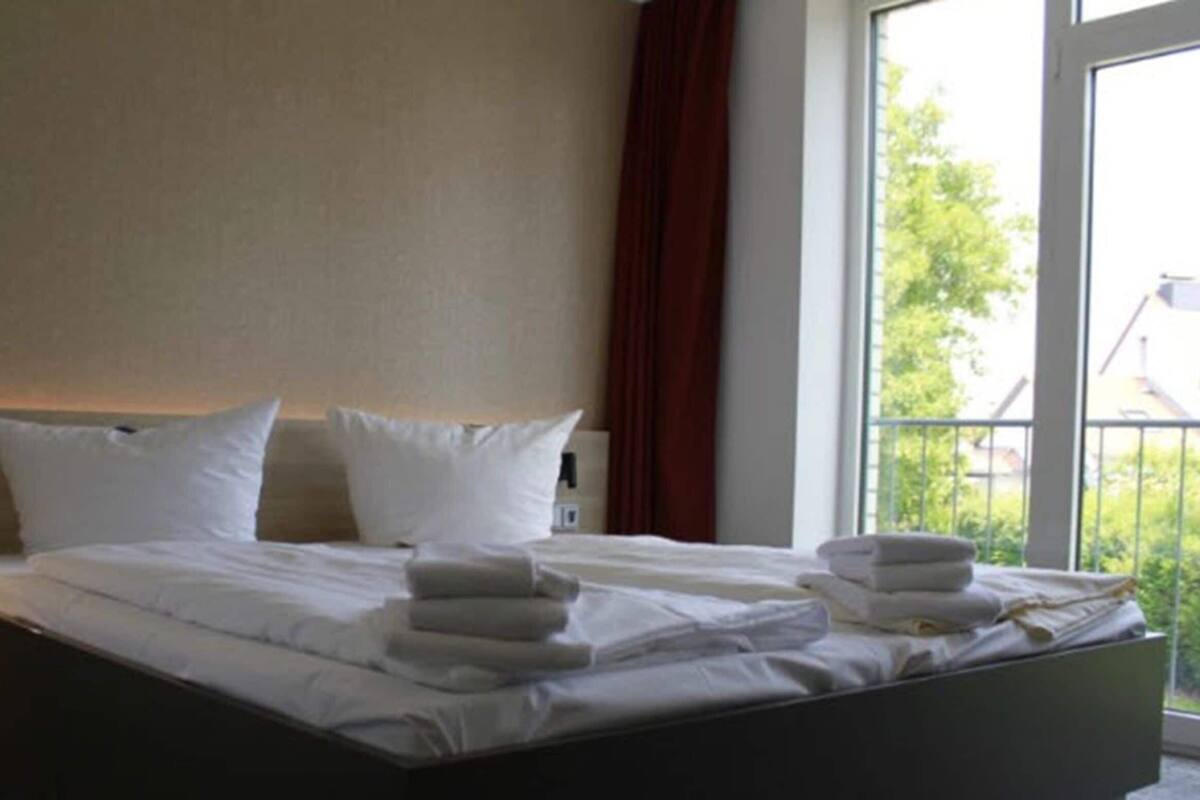 A hotel room with a bed, pillows, a nightstand, a window with a view, and a curtain.