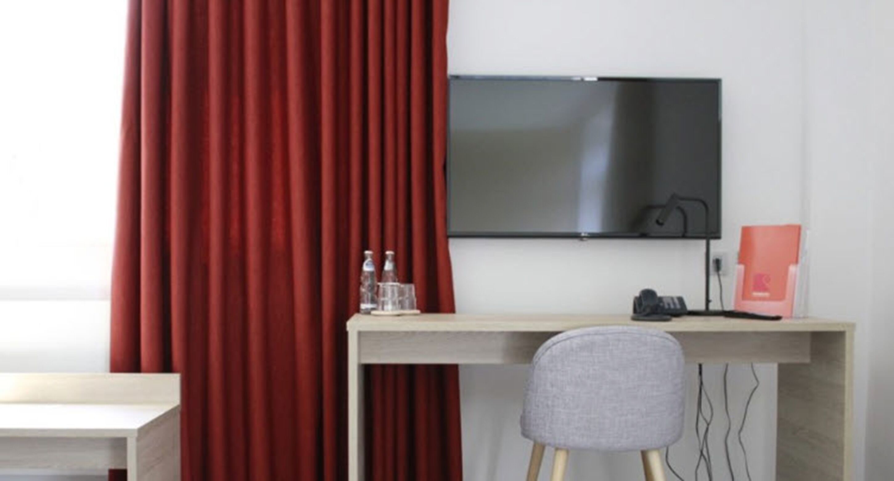 A modern room with a desk, a chair, a flat-screen TV mounted on the wall, and a red curtain.