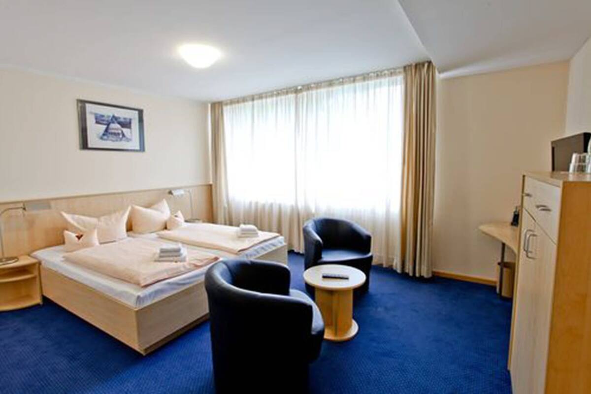 A hotel room with two beds, a desk, a chair, a small round table, and a large window with curtains.