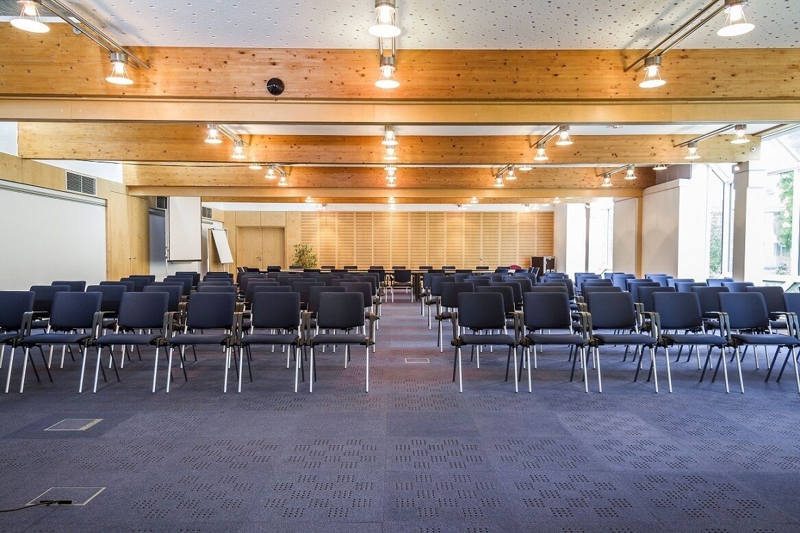 Ein Konferenzraum mit Reihen von Stühlen, Holzbalkendecke und großen Fenstern.
