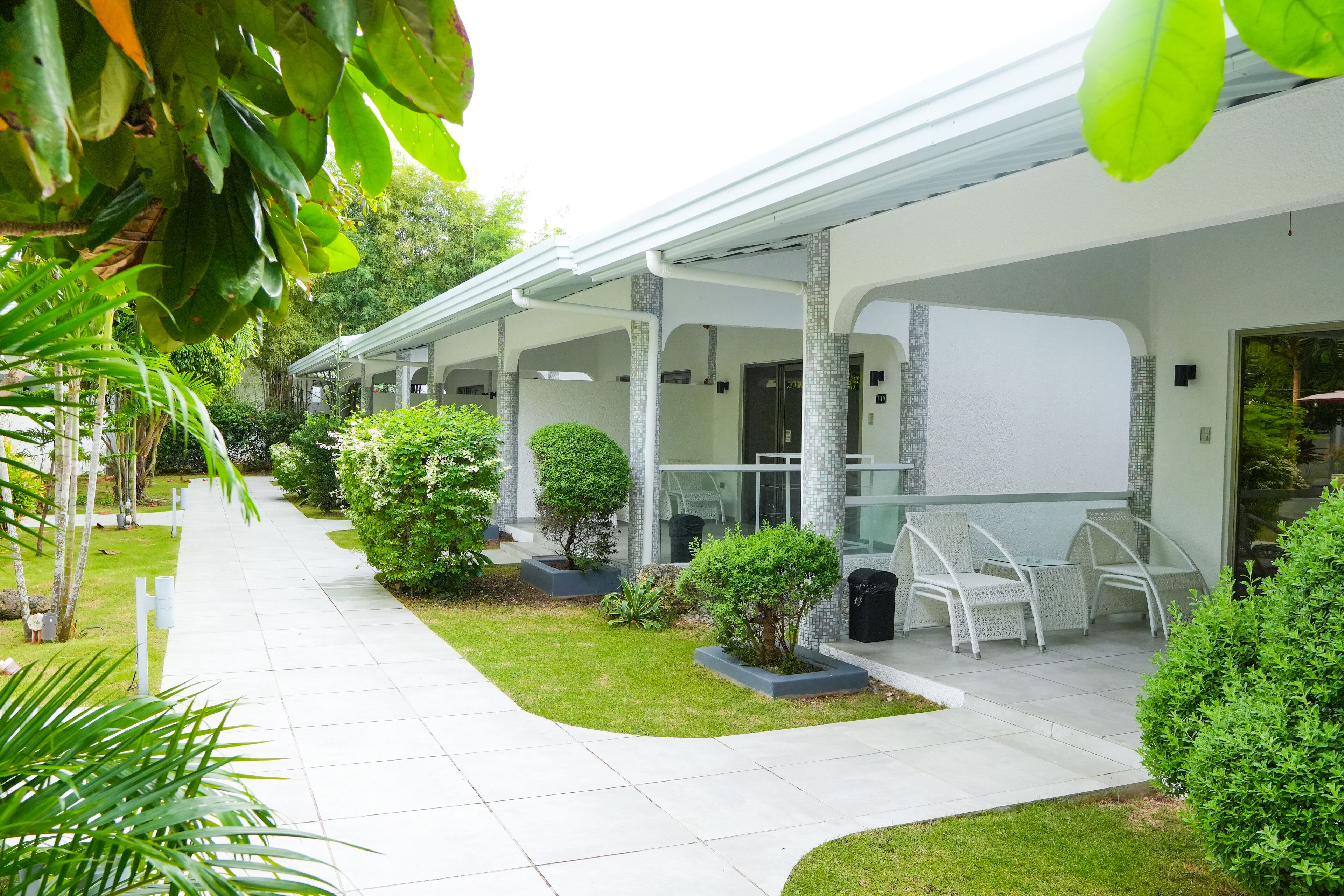 A covered outdoor seating area with white chairs and tables, surrounded by greenery and a tiled pathway.