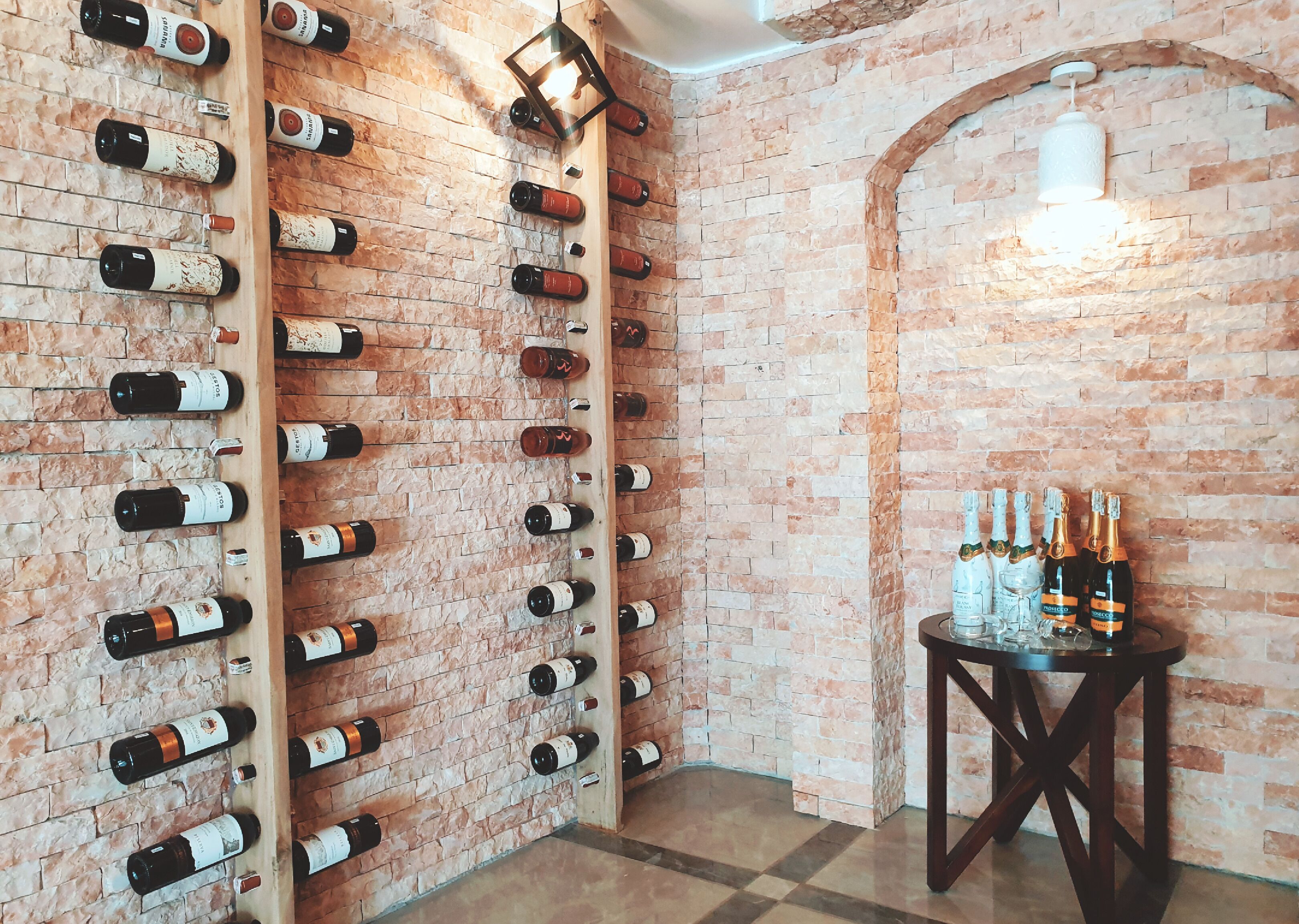 Bodega con pared de ladrillo y estanterías de madera para vino. Una pequeña mesa con botellas y copas.