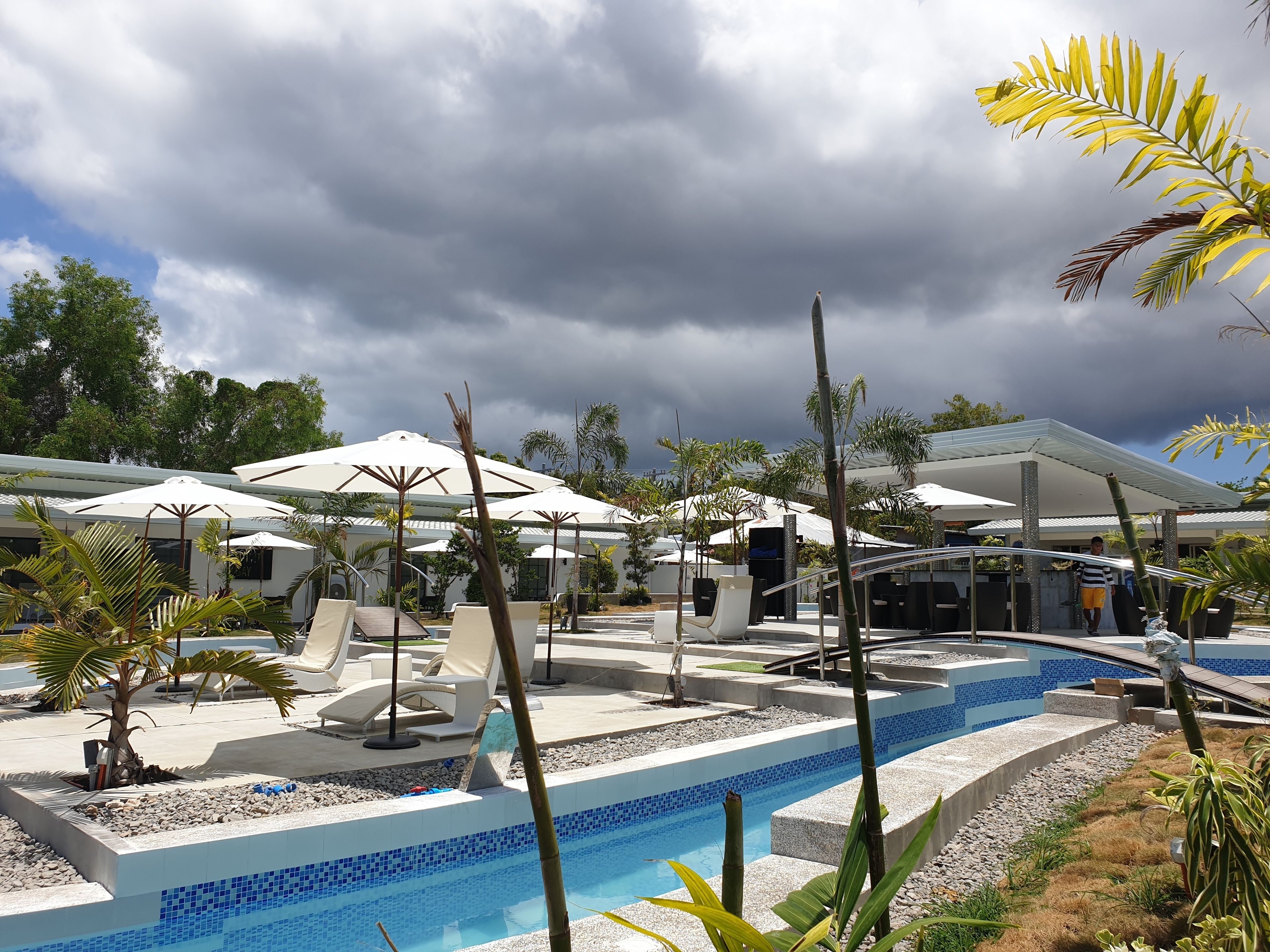 A pool area with lounge chairs and umbrellas, surrounded by palm trees and a modern building.