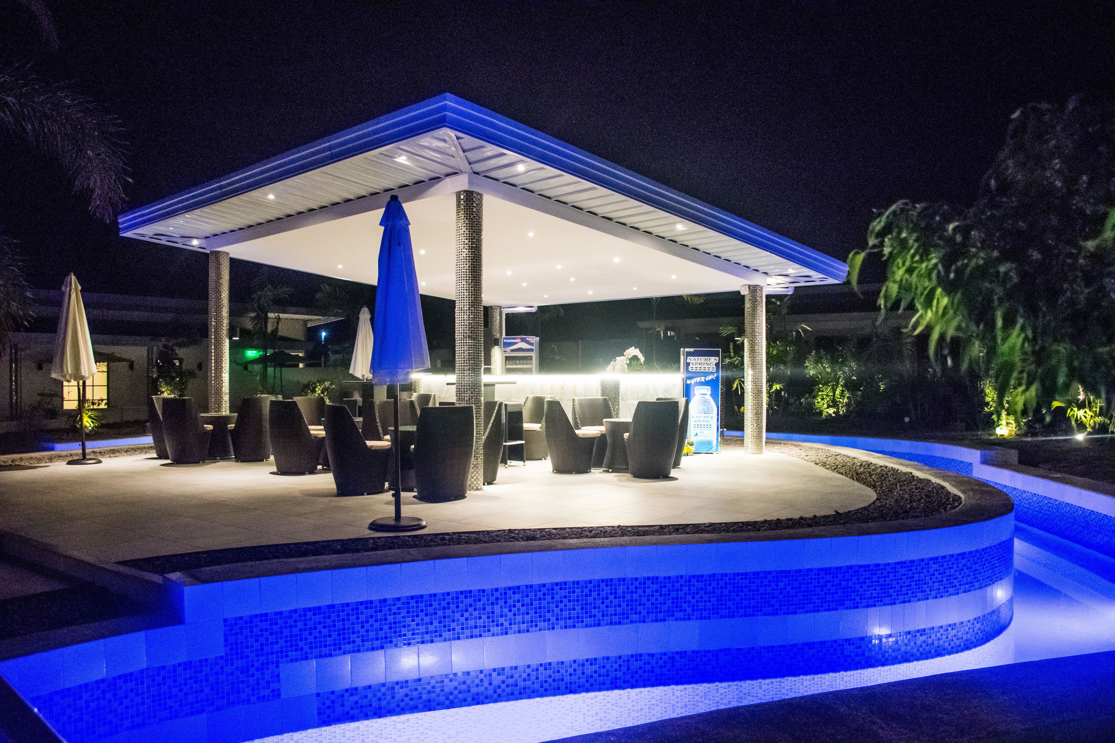 A covered outdoor seating area with tables and chairs, a pool with blue-tiled steps, and a well-lit bar area.