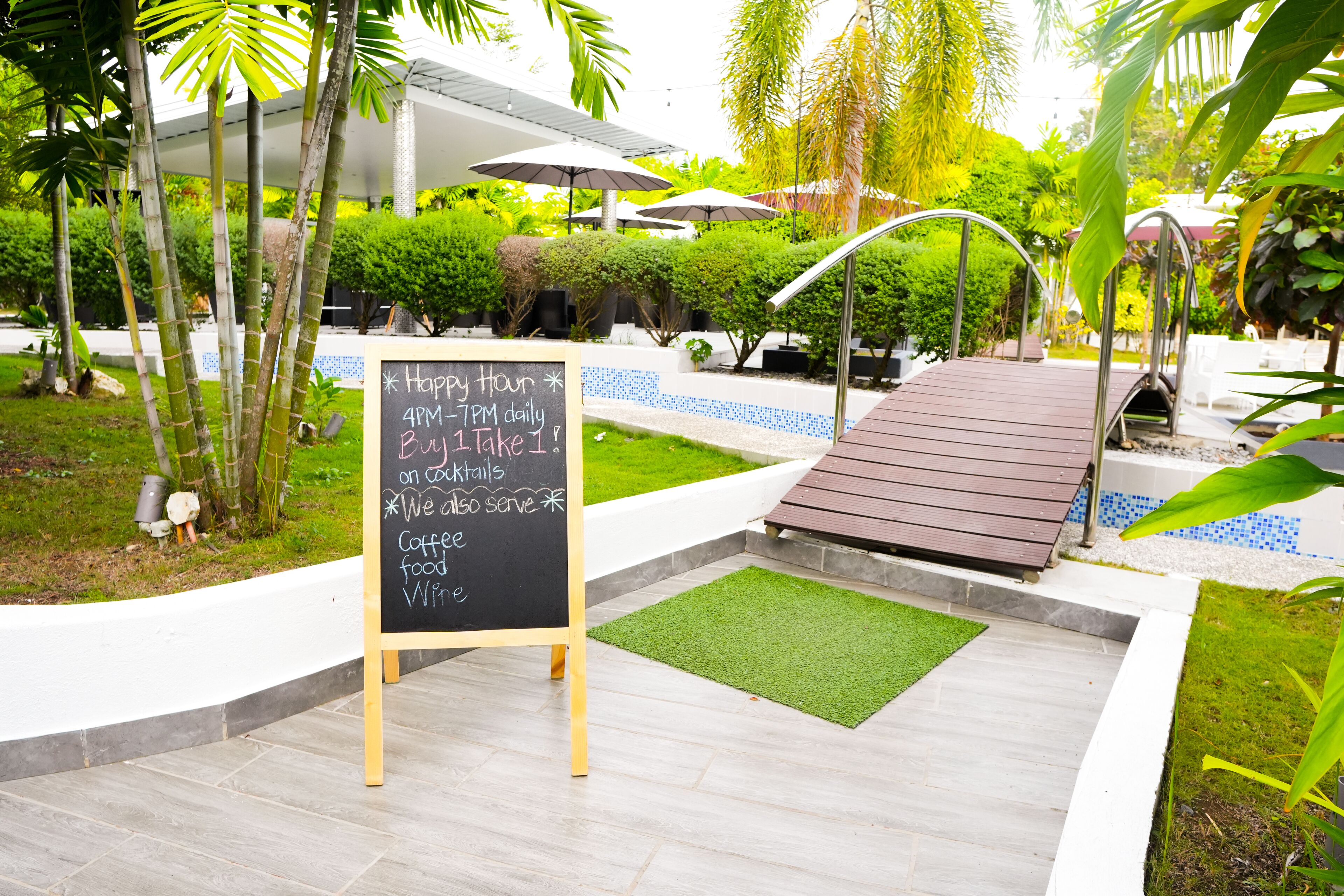 A blackboard with a happy hour schedule, surrounded by a garden with palm trees and a wooden deck.