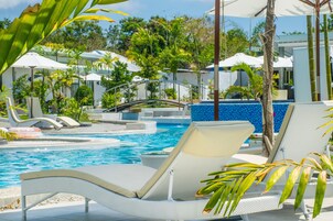 Zona de piscina con sillones de descanso y sombrillas, rodeada de plantas tropicales y una piscina azul cristalina.