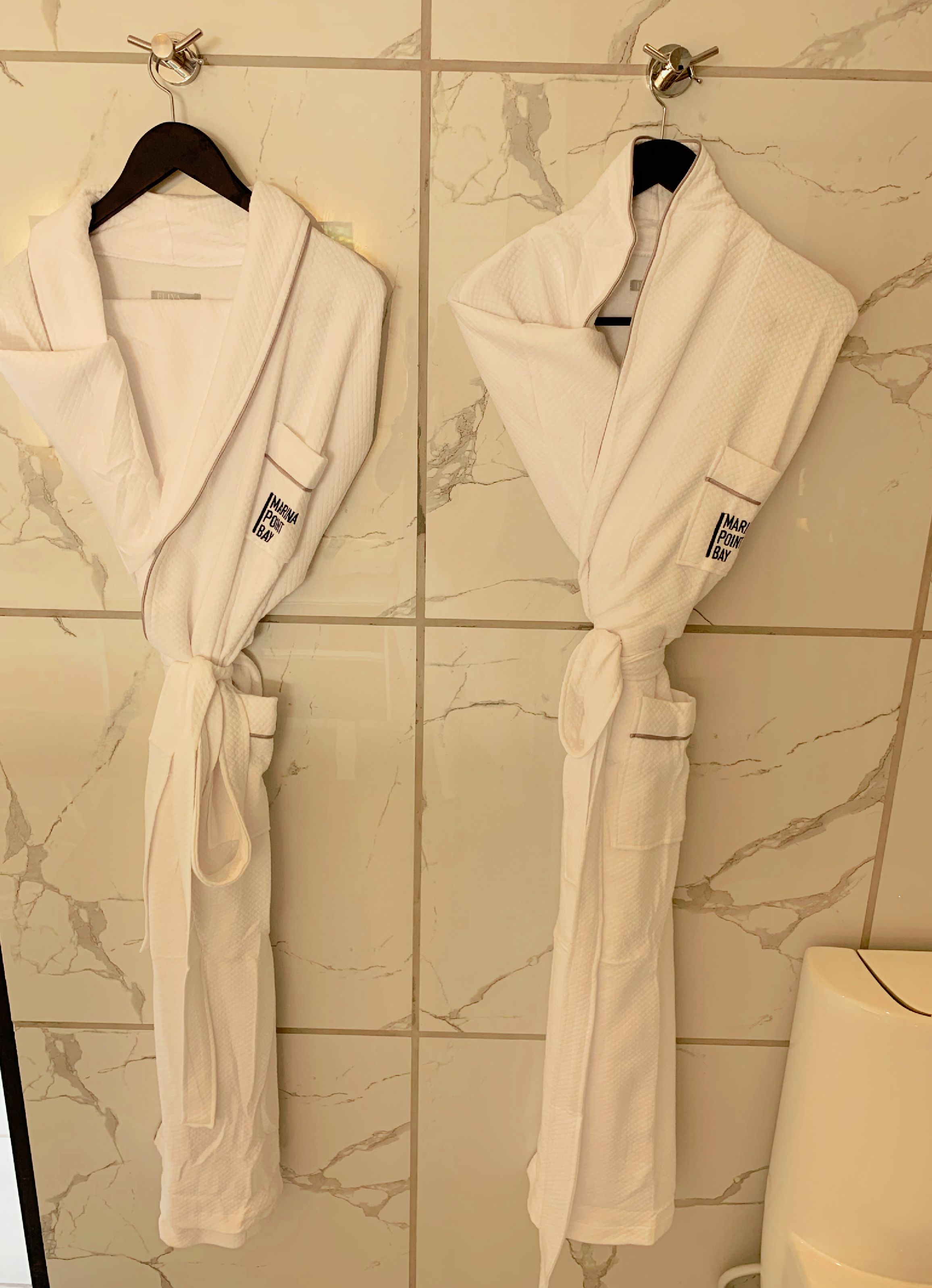 Two white robes tied to wall hooks in a bathroom with marble tiles.