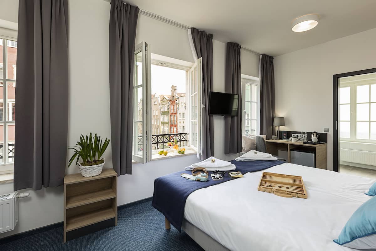 Habitación de hotel con cama, escritorio, televisión, ventana con cortinas, planta y vistas de edificios.